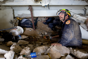 Ein Feuerwehrmann in einem Helm sitzt zwischen Trümmern, darunter Steine und Flaschen, mit einer teilweise sichtbaren Wand im Hintergrund, die wahrscheinlich den Namen des Gebäudes trägt, das von einem israelischen Luftschlag getroffen wurde.