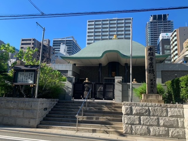 Großes grünoffices Gebäude in einem städtischen Platz mit umliegenden Treppen, Geländern, Beschilderung, Grünflächen und einer Straße darunter, vor einem klaren blauen Himmel.