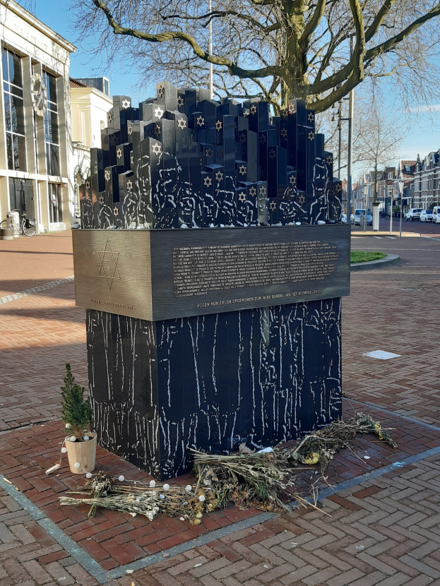 Denkmal in einem Stadtplatz für Holocaust-Opfer mit einem Baum davor, einem Topfgewächs am Boden, verstreuten trockenen Blättern und Gebäuden, Fahrzeugen, Pfählen und Bäumen im Hintergrund.