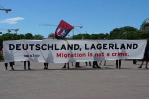 Eine Gruppe von Menschen hält ein Transparent mit der Aufschrift "Deutschland, Lagerland Migration ist kein Verbrechen" und eine Flagge, mit Straßenlaternen, Bäumen, Kränen, einem Gebäude und einem bewölkten Himmel im Hintergrund.