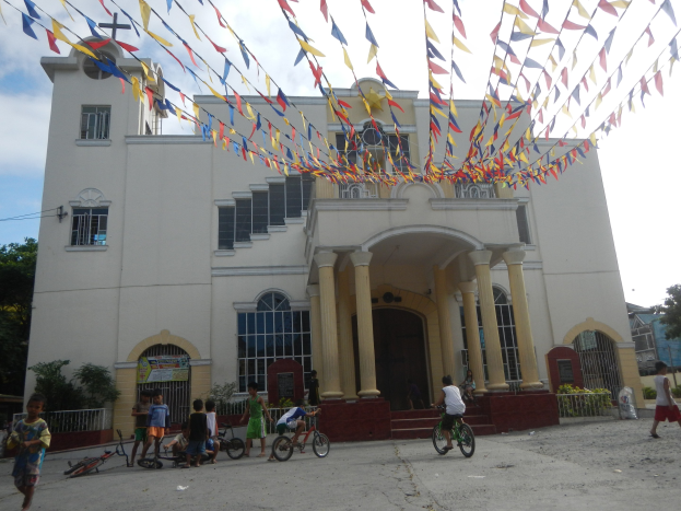 Menschen auf Fahrrädern vor einer weißen Kirche mit architektonischen Details, umgeben von Grün, einem Banner, Bäumen und bunten Fahnen vor einem bewölkten Himmel.