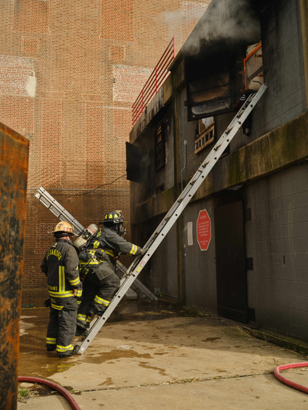 Feuerwehrleute mit Helmen klettern eine Leiter hinauf zu einem raucherfüllten Gebäude, mit einem metallenen Objekt links und Rohren an der Basis.