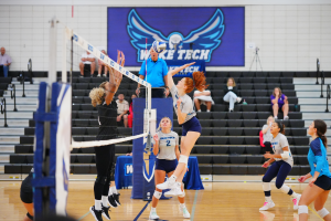 Eine Gruppe von Frauen beim Volleyballspielen auf einem Feld, mit Zuschauern auf einer Treppe im Hintergrund und einem Banner an der Wand, das einen Adler und einen Text zeigt, der den Sieg der Frauen-Volleyballmannschaft bei der Staatsmeisterschaft feiert.