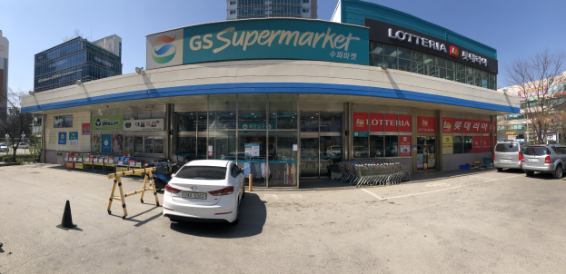 Ein Auto vor einem "GS Supermarkt" parkend mit anderen Fahrzeugen, Verkehrskegeln und verstreuten Gegenständen auf der Straße, vor Häusern, Bäumen und einem klaren blauen Himmel.