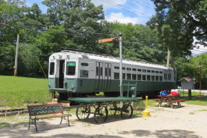Ein Zug auf Schienen mit einer Bank und einem Tisch in der Nähe, umgeben von Gras, Bäumen, Drähten und einem Pfahl unter einem klaren Himmel.