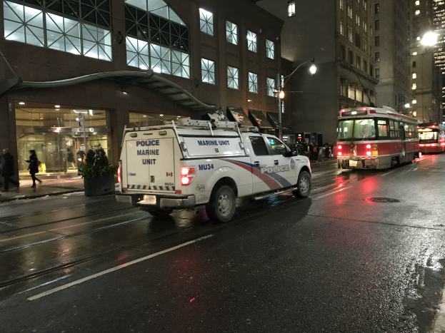Ein Polizei-Lkw parkt nachts am Straßenrand, mit Fahrzeugen auf der Straße, Fußgängern auf dem Gehweg, Topfpflanzen, Straßenlaternen, Gebäuden mit Fenstern und einer bewölkten Himmel.