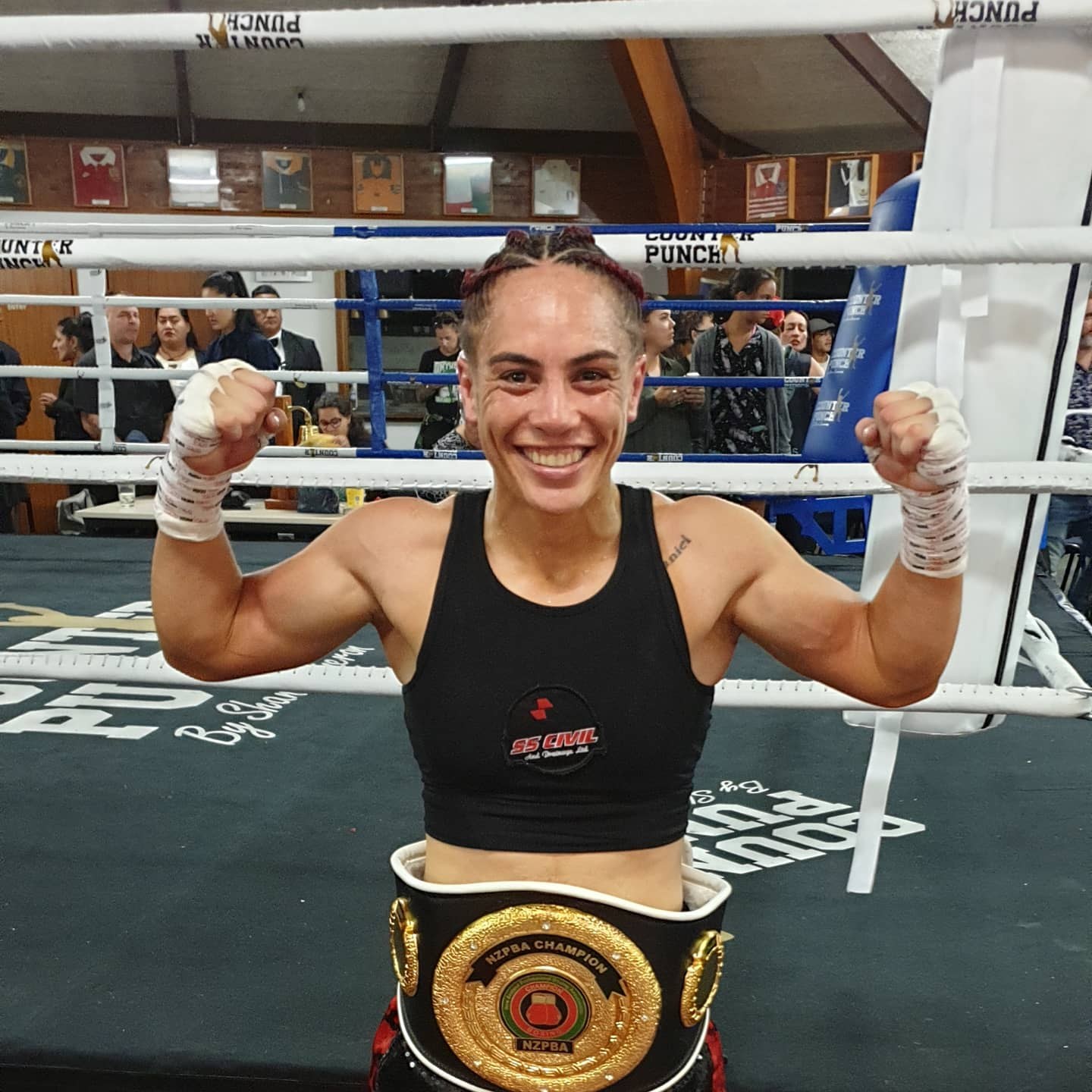 Eine Frau in einem schwarzen Kleid und einem goldenen Gürtel steht in einem Boxring mit erhobenen Händen, feiernd als Junior-Weltergewicht-Champion, mit Zuschauern und Tafeln im Hintergrund.
