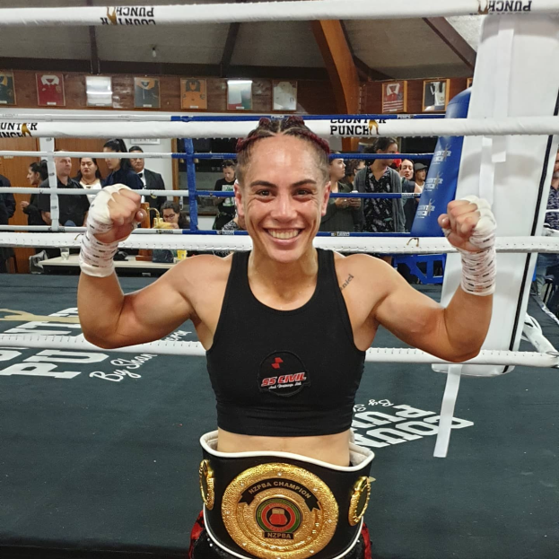 Eine Frau in einem schwarzen Kleid und einem goldenen Gürtel steht in einem Boxring mit erhobenen Händen, feiernd als Junior-Weltergewicht-Champion, mit Zuschauern und Tafeln im Hintergrund.