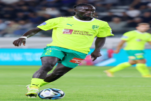 Ein Fußballspieler in einem gelben T-Shirt und grünen Shorts ist im Einsatz auf dem Feld, mit einer blauen Tafel und unscharfen Menschen im Hintergrund.