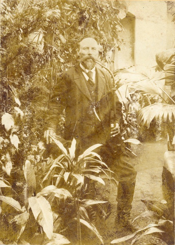 Altes Foto eines Mannes in Anzug und Krawatte, der in einem Garten mit Pflanzen, Bäumen und einer Wand im Hintergrund steht.