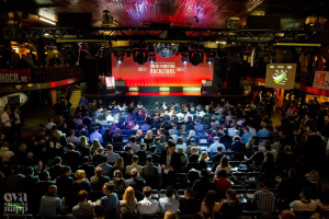 Eine große Menschenmenge sitzt auf Stühlen vor einer Bühne mit Bildschirmen und Bannern, beleuchtet von Scheinwerfern und umgeben von Geländern, mit einem Wasserzeichen in der linken unteren Ecke.