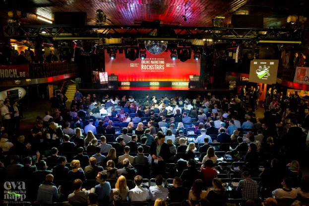Eine große Menschenmenge sitzt auf Stühlen vor einer Bühne mit Bildschirmen und Bannern, beleuchtet von Scheinwerfern und umgeben von Geländern, mit einem Wasserzeichen in der linken unteren Ecke.