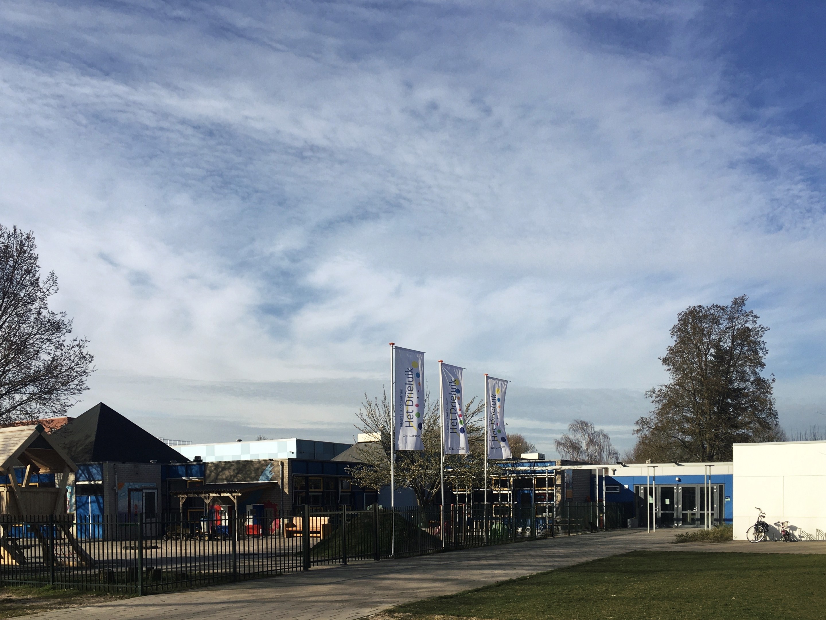Großes Gebäude mit Metallzaun, Rasenfläche, Weg, Bäumen, Bannern, einem geparkten Fahrrad, nahen Häusern und einem bewölkten Himmel, identifiziert als der neue Spielplatz der Schule.