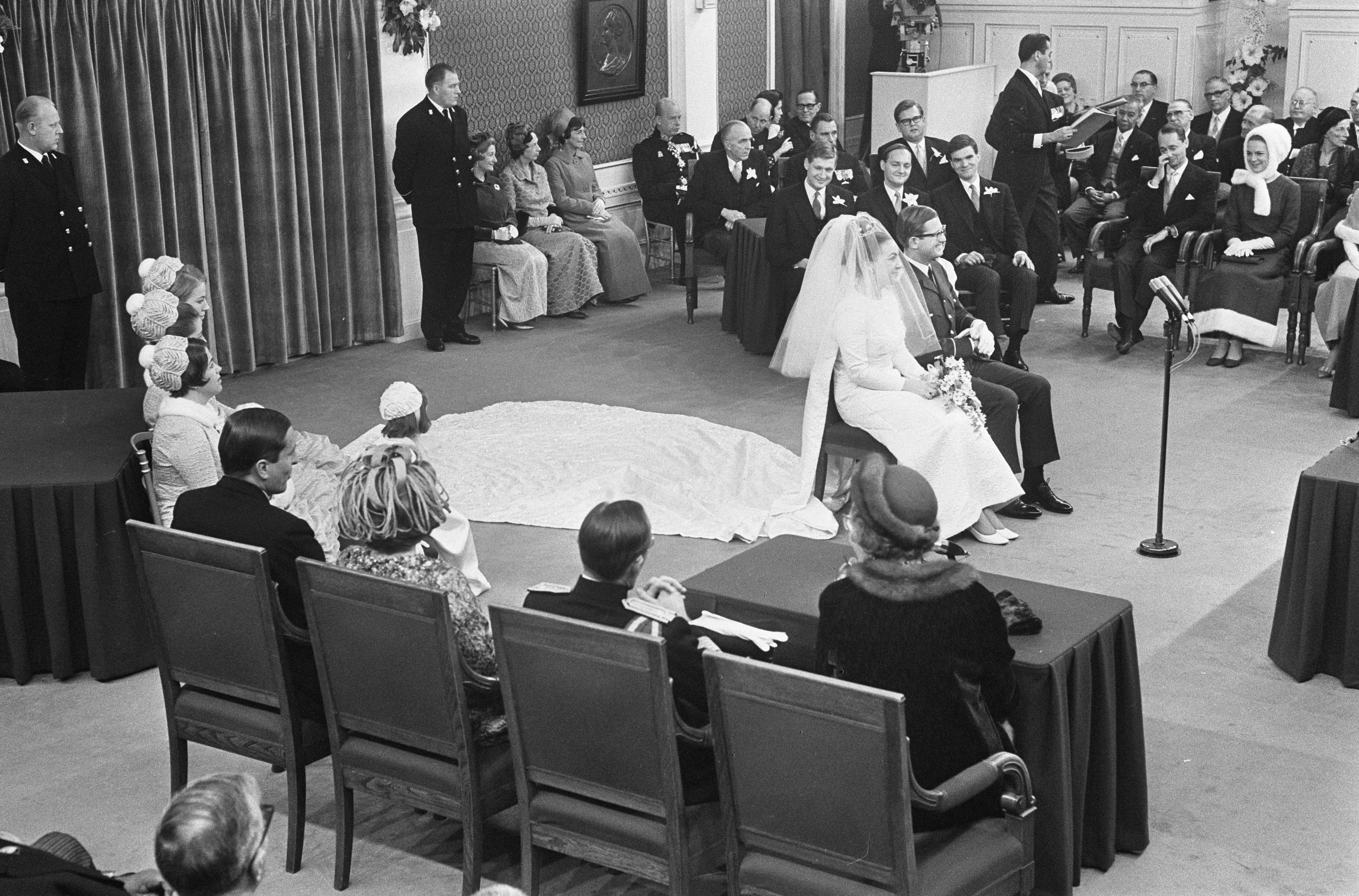 Schwarzes und weißes Foto einer Hochzeitszeremonie mit sitzenden und stehenden Menschen, ein Mann mit Mikrofon, eine Frau mit einem Blumenstrauß, ein Podium mit Mikrofon, Blumengestecke, ein Vorhang, ein gerahmtes Foto an der Wand und eine Tür im Hintergrund.