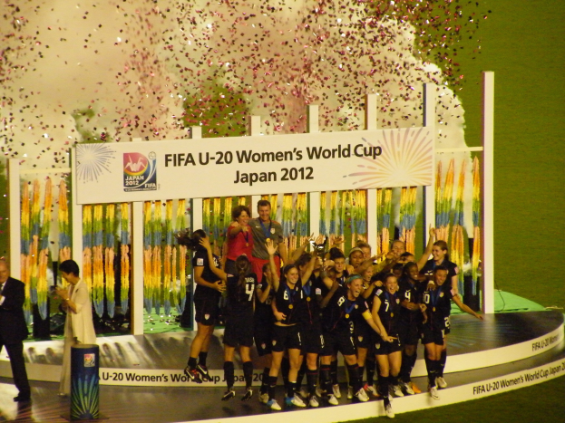 Gruppe von Frauenfußballspielerinnen in schwarzen Uniformen, die auf einer Bühne umgeben von einer Menge feiern, mit einer Tafel und dekorativen Elementen im Hintergrund.