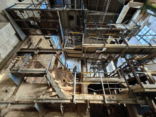 Altes Industriegebäude mit einem zentralen Metallgerüst, verrosteten Wänden und verfallendem Boden, umgeben von Eisenstangen und Treppen.