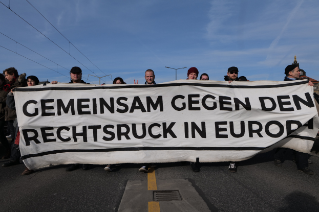 Eine Gruppe von Menschen steht auf der Straße und hält ein Transparent mit der Aufschrift "Gemeinsam gegen den Rechtsruck in Europa" mit Straßenlaternen, Strommasten, Stromkabeln, Gebäuden und einem bewölkten Himmel im Hintergrund.