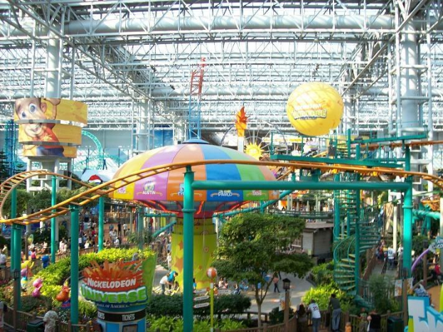 Großer Indoor-Wasserpark mit Achterbahn, umgeben von Menschen, Bäumen, Pflanzen und Deckenlampen.