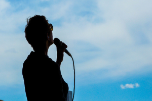 Eine Person mit Brille hält ein Mikrofon vor einem Himmel-Hintergrund.