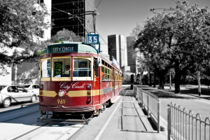 Ein roter Zug mit einer Person darin und Text darauf ist in der Mitte, mit Autos, Bäumen, Gebäuden, Laternenmasten und Drähten links und Bäumen und einem weißen Zaun rechts, mit Gebäuden und einer Person im Hintergrund.