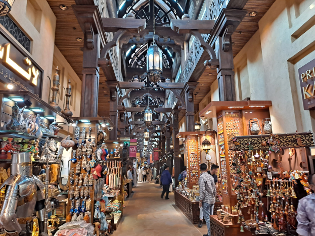 Ein geschäftiges Dubai-Bazar mit Menschen, die zwischen Regalen voller Gegenstände hindurchgehen, Wänden mit Texttafeln und einer Decke mit warmem Licht.