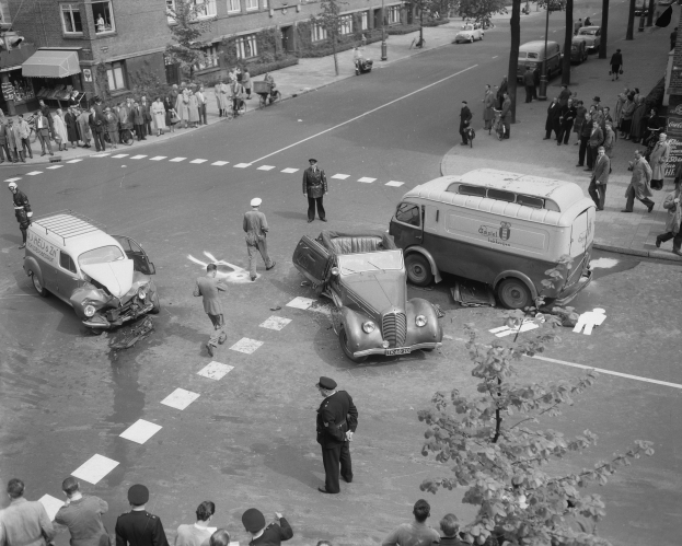 Schwarz-weißes Bild einer Autounfall-Szene an einer Straßenkreuzung mit mehreren Fahrzeugen, darunter ein Lieferwagen, und einer Gruppe von Schaulustigen; Gebäude, Bäume, Pfosten und Schilder sind im Hintergrund zu sehen.