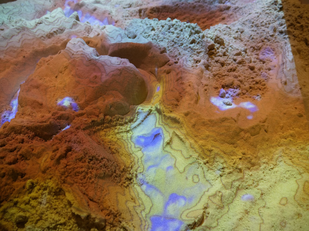 Nahaufnahme einer detaillierten 3D-Karte einer Wüstenlandschaft mit sandfarbenen Bergen, Tälern und Graten, beleuchtet von hellem Licht.