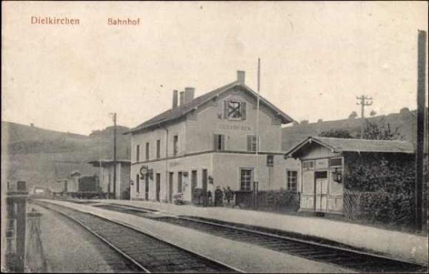 Schwarzes und weißes Foto einer Bahnstation in Dielkirchen, Deutschland, mit einem Zug auf den Gleisen, umgeben von Häusern, Strommasten, Bäumen, Menschen und Hügeln im Hintergrund bei klarem Himmel.
