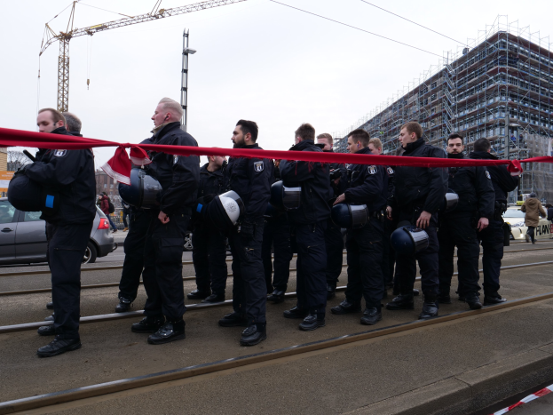 Eine Gruppe von Polizisten in schwarzen Uniformen und Helmen steht in einer Reihe auf einem Bahngeleis mit einem roten Band davor, wobei Fahrzeuge, Passanten, Gebäude und Versorgungsmäste im Hintergrund unter einem klaren blauen Himmel zu sehen sind.