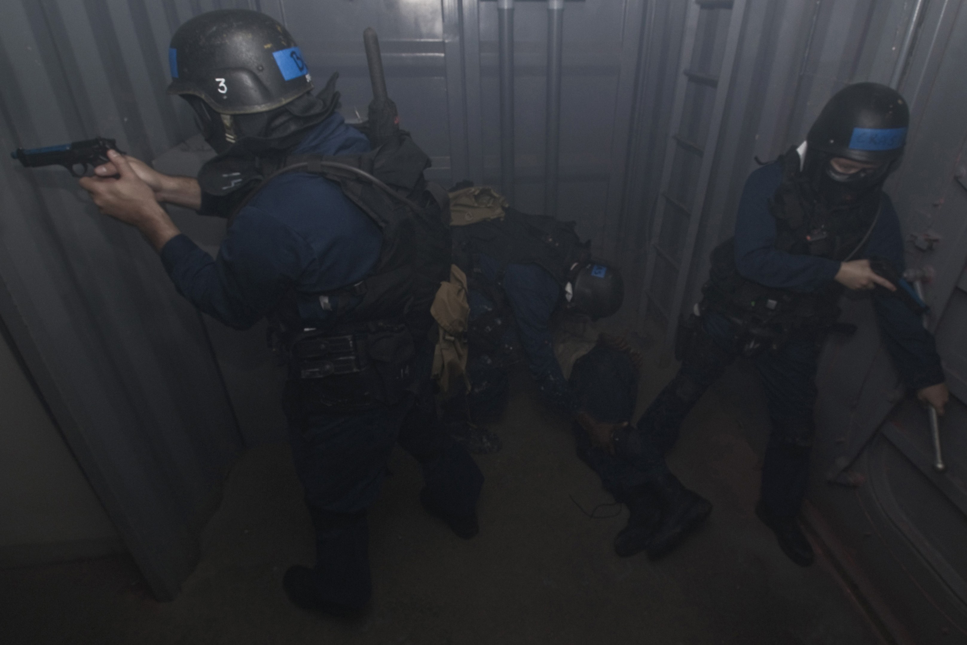 Polizisten in Einsatzkleidung, einer mit einer Waffe, neben einem Metallcontainer und einer Leiter in einem beengten Raum.