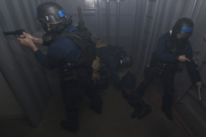 Polizisten in Einsatzkleidung, einer mit einer Waffe, neben einem Metallcontainer und einer Leiter in einem beengten Raum.