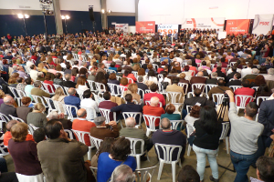 Ein volles Stadion mit vielen sitzenden und stehenden Menschen, eine Bühne mit zwei Rednern auf einem Podium und zahlreiche Banner und Lichter.