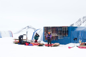 Drei Personen stehen auf einer schneebedeckten Landschaft mit verstreuten Taschen, Zelten mit Skiern dahinter und schneebedeckten Hühgeln im Hintergrund unter einem klaren Himmel.