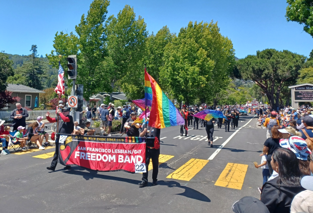 Eine Gruppe von Menschen, die auf der Straße gehen, sitzen und stehen, während sie Regenschirme halten und eine Regenbogenflagge tragen, mit einer Ampel, einem Schild, Bäumen und Gebäuden im Hintergrund.