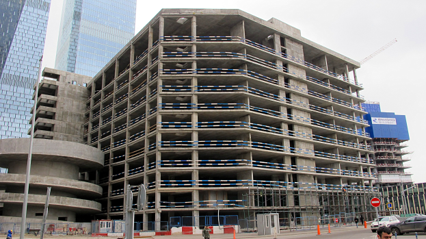 Großer Bau in einer städtischen Umgebung mit Wolkenkratzern, Fahrzeugen, Fußgängern, Verkehrs-Infrastruktur und Kränen vor einem klaren Himmel.