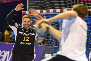 Zwei Männer spielen Handball vor einem Tor mit Netz und einem Banner mit Text im Hintergrund und einigen Zuschauern in einem Stadion.