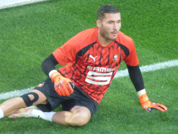 Ein Fussballspieler in rotem T-Shirt, schwarzen Shorts, weissen Socken und orangen Baseball-Handschuhen sitzt auf dem Boden mit Händen auf den Knien.