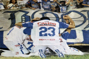 Gruppe von Fußballspielern, die auf dem Boden vor einer jubelnden Menge feiern, mit einem Banner mit Text im Hintergrund.