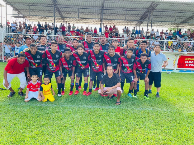 Mannergruppe feiert auf einem Fußballfeld mit einem Siegesschild, das "Fotos Ugarte U-17 vs U-18" trägt, unter einem bewölkten Himmel.