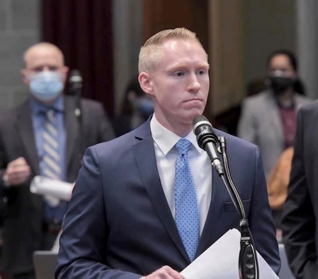 Ein Mann in Anzug und Krawatte spricht an einem Podium mit Papieren vor einem Mikrofon, mit ein paar Leuten und einer Vorhangwand im Hintergrund.