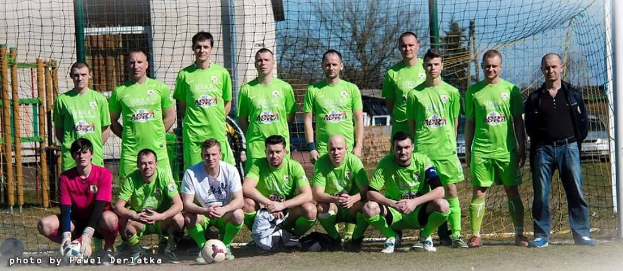 Eine Gruppe von Männern in grünen T-Shirts steht oder hockt auf einem Fußballfeld in der Nähe eines Torpfostens mit einem Ball davor, mit Bäumen, Gebäuden und Fahrzeugen im Hintergrund.