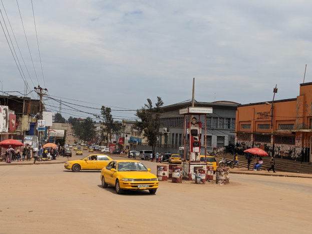 Ein gelbes Taxi fährt eine belebte Stadtstraße entlang, die von Gebäuden gesäumt ist, während Fußgänger mit Schirmen auf dem Gehweg unter einem bewölkten Himmel gehen.