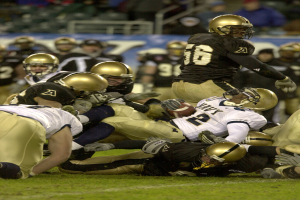 Eine Gruppe von Menschen in Helmen beim Fußballspielen auf einem Feld, im Hintergrund sind Menschen, Geländer und eine Texttafel zu sehen.