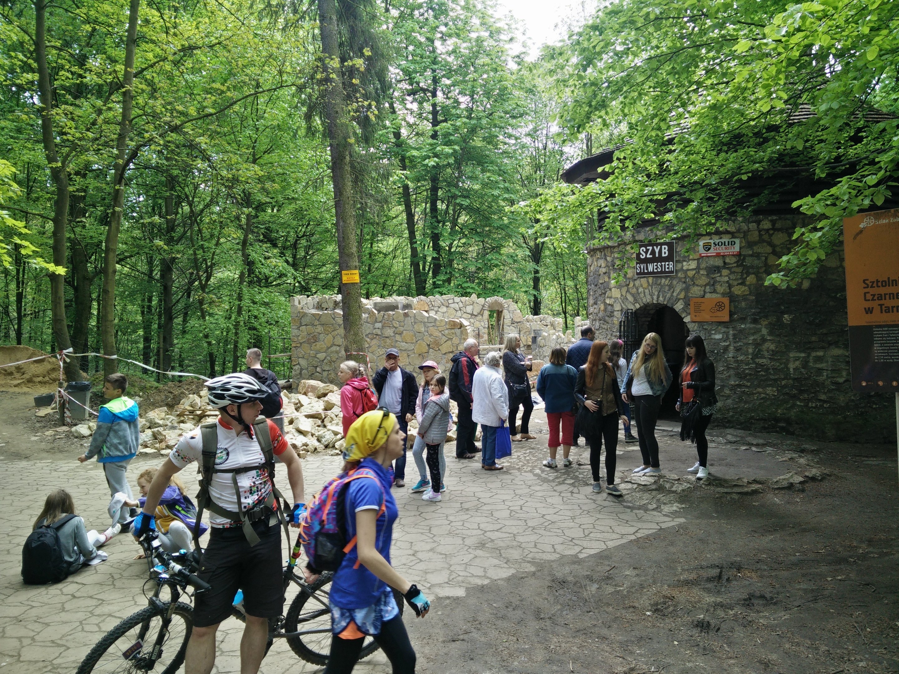 Gruppe von Menschen, die einen bewaldeten Pfad entlanggehen, einige tragen Helme und tragen Taschen oder Fahrräder, mit einer Steinmauer und einem Schild im Hintergrund, das den Eingang zum Szyb Stylweste Mountain Bike Trail anzeigt.
