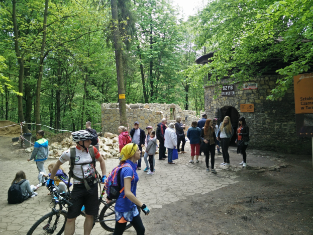 Gruppe von Menschen, die einen bewaldeten Pfad entlanggehen, einige tragen Helme und tragen Taschen oder Fahrräder, mit einer Steinmauer und einem Schild im Hintergrund, das den Eingang zum Szyb Stylweste Mountain Bike Trail anzeigt.