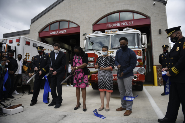 Gruppe von Menschen mit blauen Bändern vor einem Feuerwehrauto bei einer Einweihungsfeier für eine neue Feuerwache, mit einem Gebäude und Himmel im Hintergrund.