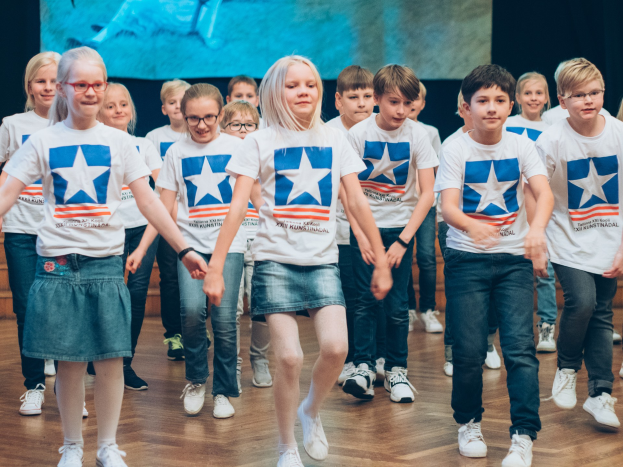 Gruppe von Kindern in weißen Hemden und blauen Shorts tanzt auf einem Holzfußboden während eines Kunstnädler-Wettbewerbs.