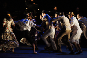 Eine Gruppe von Menschen tanzt mit anderen und Equipment im Hintergrund.