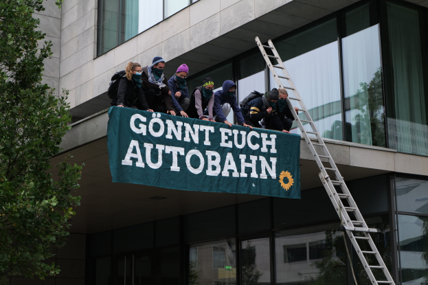 Eine Gruppe von Menschen auf dem Dach eines Gebäudes, die ein Transparent halten, mit einem Baum links und Glasfenstern im Hintergrund, wahrscheinlich protestierend für die deutsche Regierung.