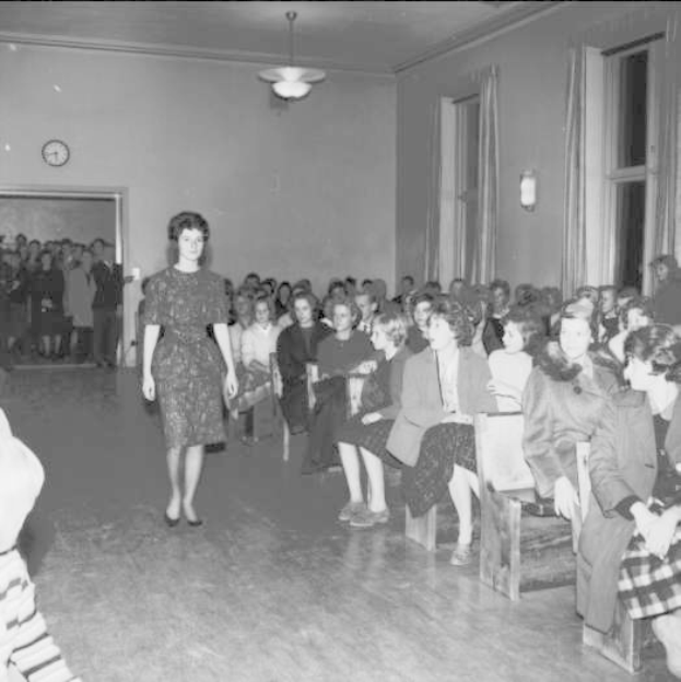 Eine Frau in einem schwarzen und weißen Kleid geht über einen Laufsteg bei einer Modenschau für Frauenrechte, mit einer Sitzreihe und einer Uhr und Lichtern an der umgebenden Wand.